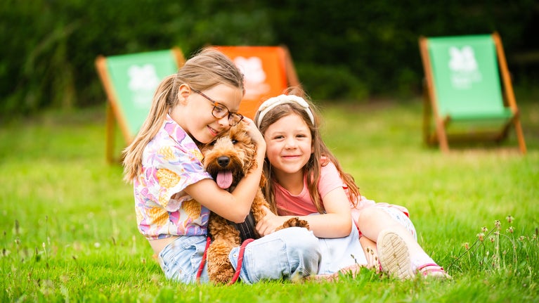 Two young girls hugging a brown cockerpoo dog whilst sitting on the grass. Deckchairs can be seen in the background